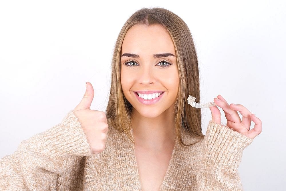 A brunette handsigning thumbs up while holding Invisalign clear aligners from Wink Orthodontics, an orthodontist in Prosper, TX