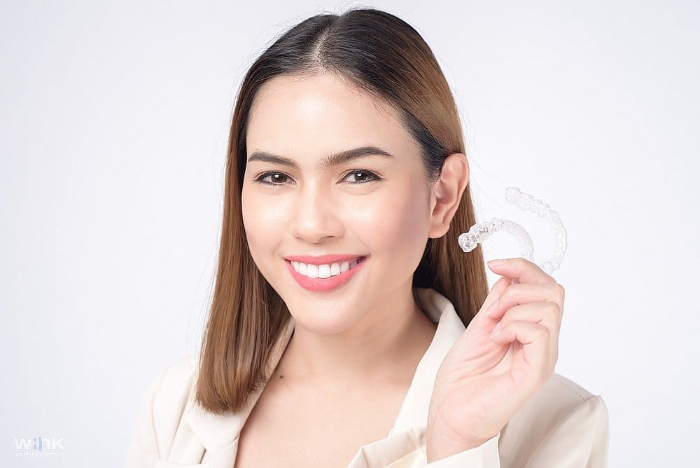 A smiling woman holding a clear aligner, showcasing an effective solution for how to fix overbite in Prosper and Colleyville TX.