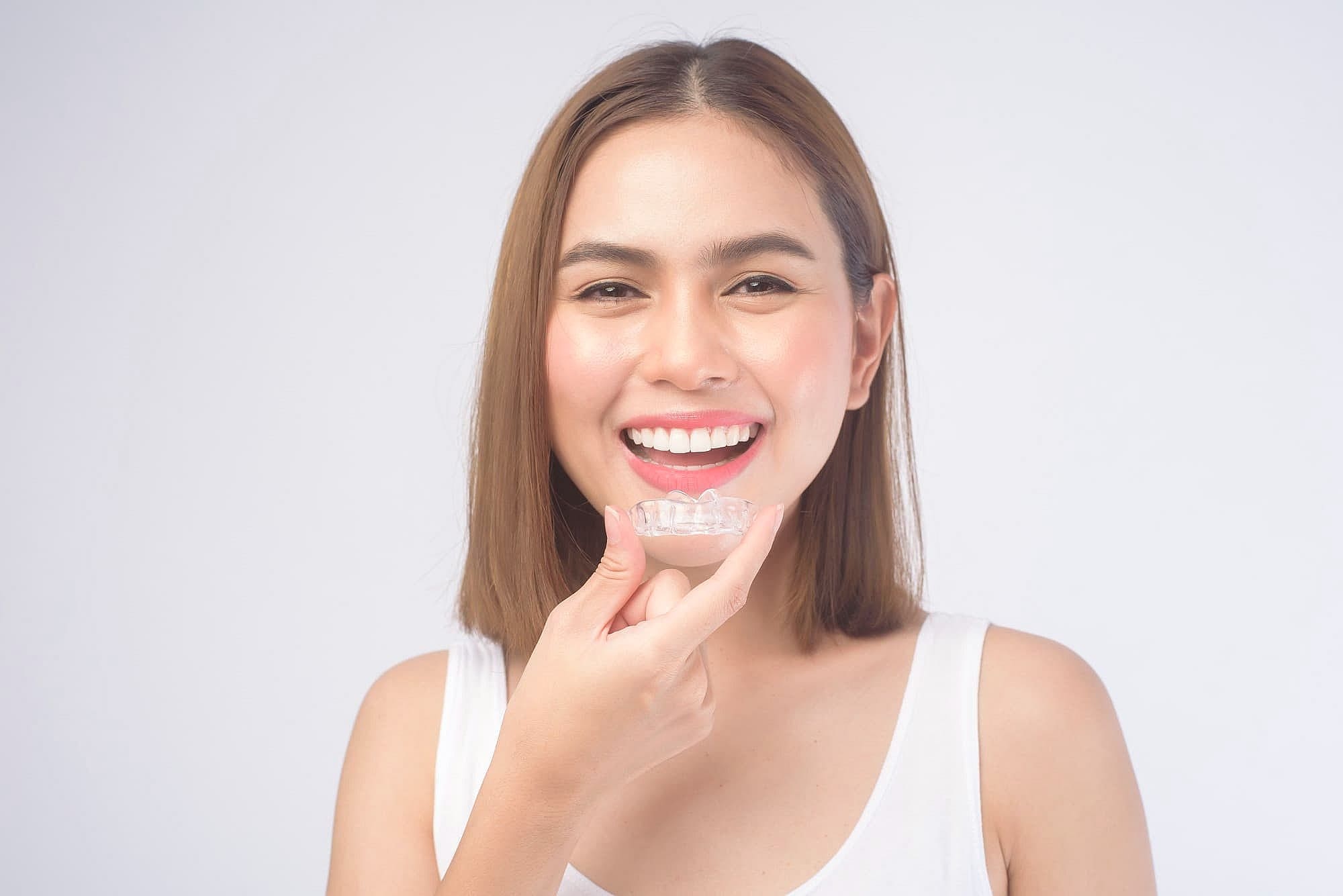A woman in a white tank top smiles while holding Invisalign® Clear Aligners near her mouth against a plain white background at Wink Orthodontics in Colleyville, TX.