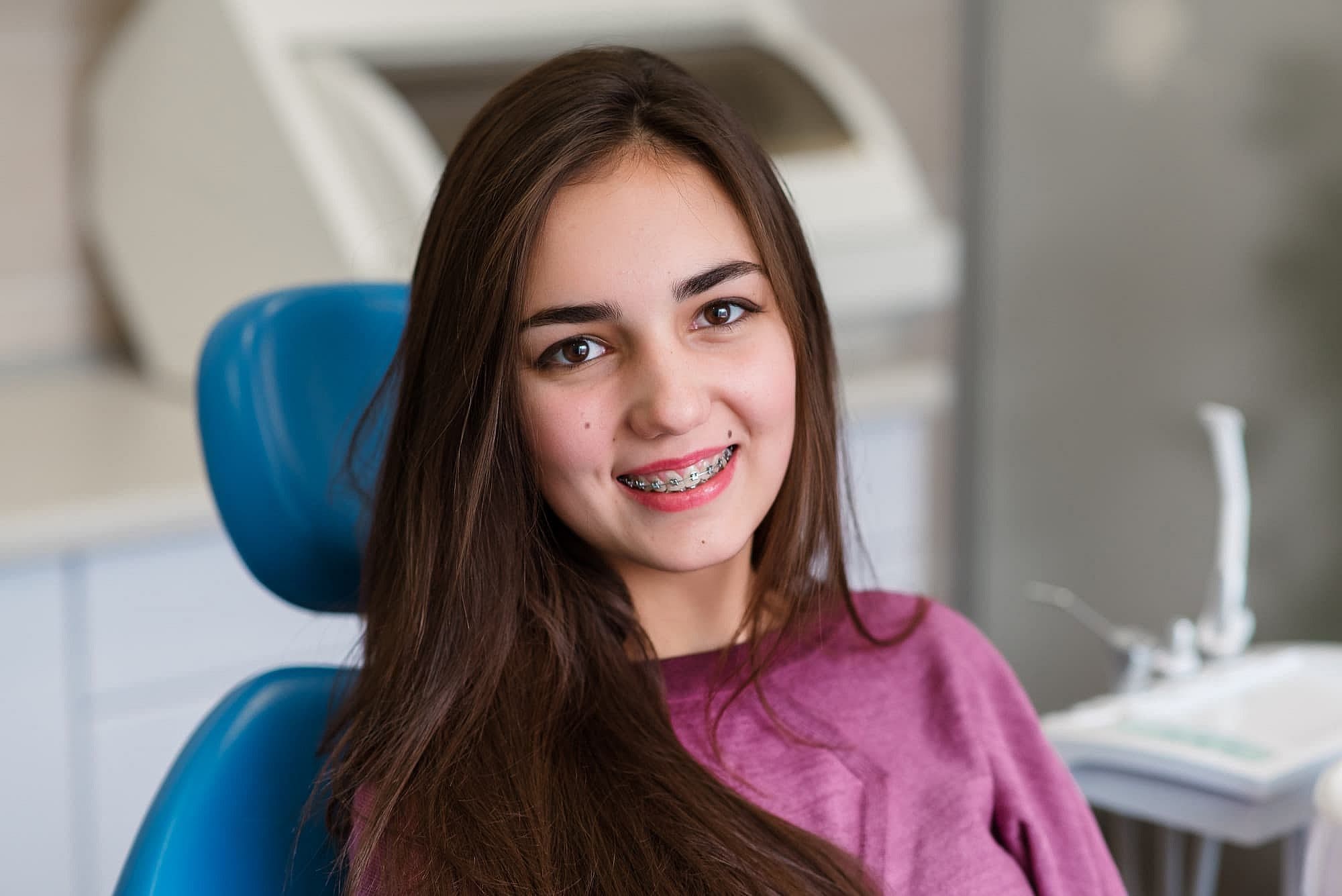A young woman with long brown hair wearing lingual braces and a mauve sweatshirt smiles in a dental chair at Wink Orthodontics in Prosper, TX, with dental equipment visible in the background.