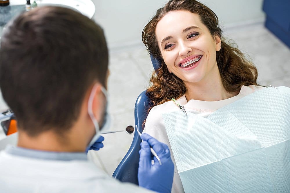 At Wink Orthodontics in Prosper, TX, a young woman with braces smiles in the dental chair as she looks at her dentist during an orthodontic checkup.