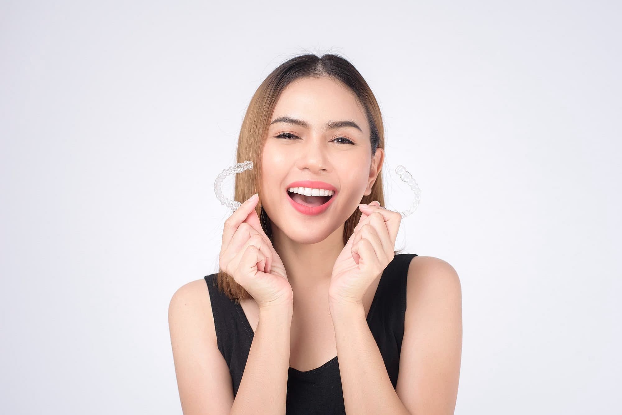 A woman in a black top smiles while holding Invisalign® Clear Aligners near her face, pictured against a plain white background for Wink Orthodontics in Prosper, TX.