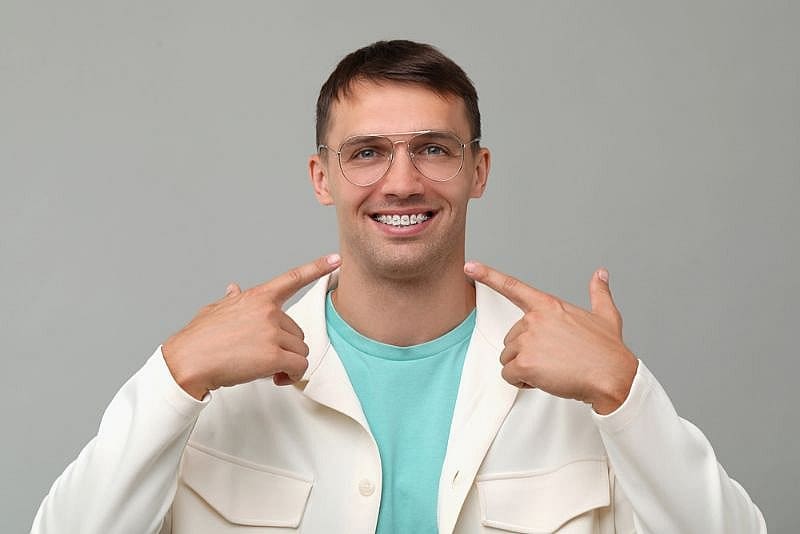 At Wink Orthodontics in Colleyville, TX, a man in glasses and a white jacket stands against a plain gray background, smiling and proudly pointing to his braces with both hands.