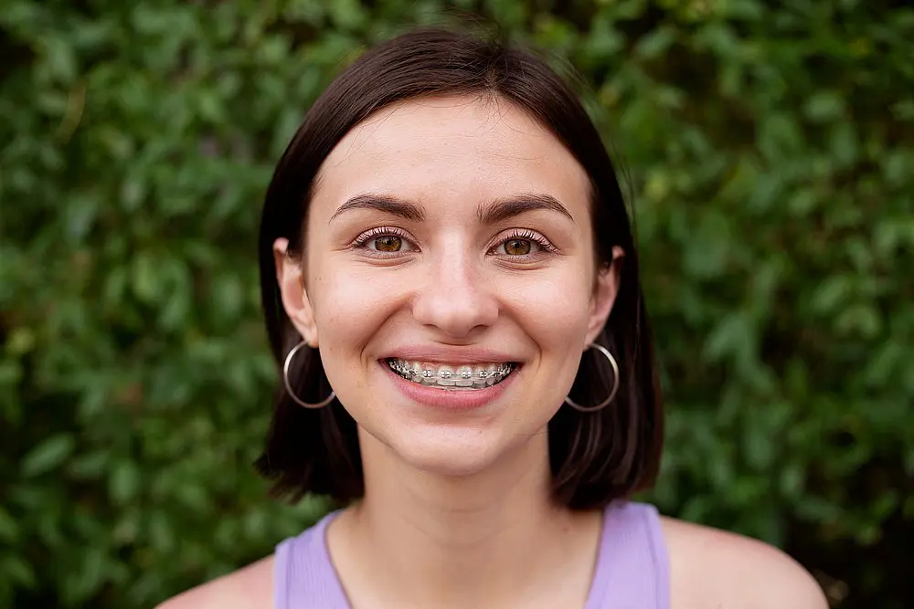 A young woman with straight brown hair, hoop earrings, and metal braces smiles at the camera against a leafy green backdrop, reflecting the cheerful atmosphere at Wink Orthodontics in Colleyville, TX.