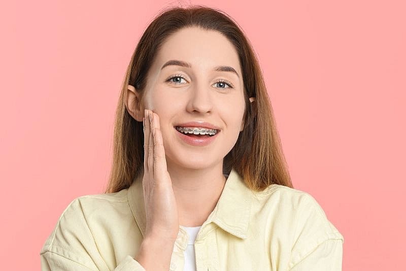 A young woman with long brown hair and stylish braces smiles, touching her cheek as she stands before a pink background—showcasing the confident results from Wink Orthodontics in Prosper, TX.