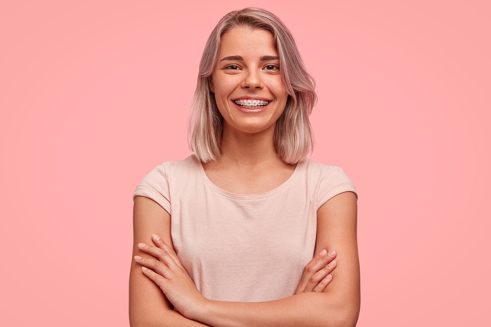 A young, light-haired woman in a light pink t-shirt confidently smiles with braces and crossed arms against a solid pink background—showcasing the results from Wink Orthodontics in Prosper, TX.