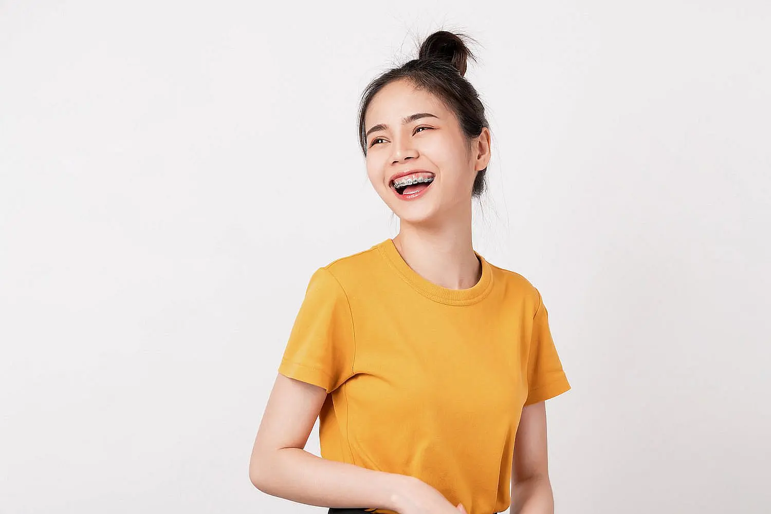 A young woman with braces, dressed in a yellow t-shirt, smiles confidently and glances to the side against a white background—showcasing results you can achieve at Wink Orthodontics in Colleyville, TX.