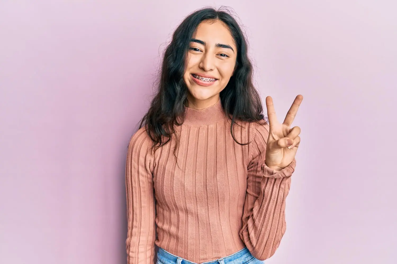 Smiling teen girl with metal braces flashes a peace sign, promoting Wink Orthodontics in Prosper or Colleyville, TX, on pink background.