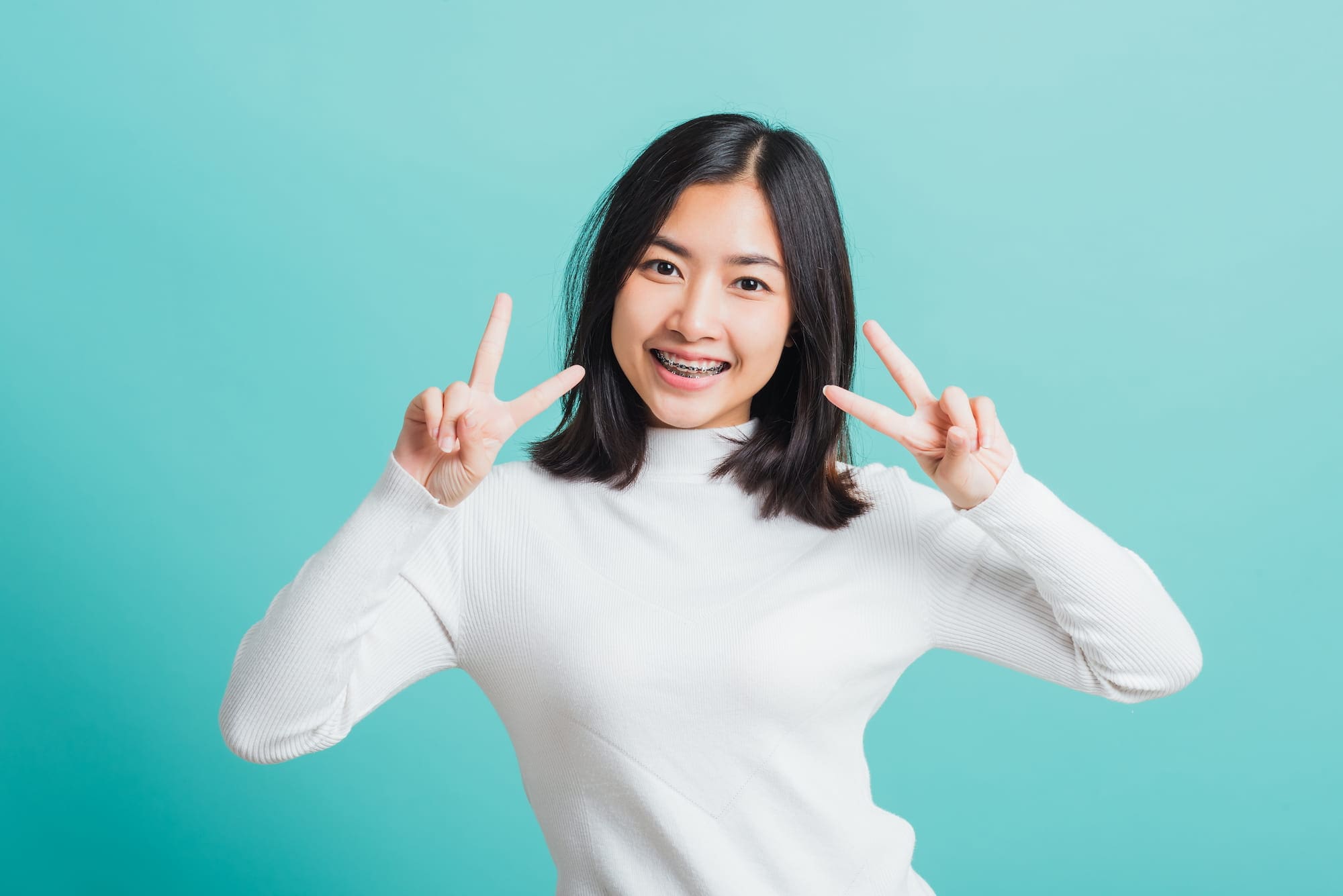 Smiling young woman with braces and overbite makes peace signs, wearing white top at Wink Orthodontics in Prosper or Colleyville, TX.