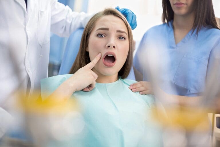 Woman sitting in a dental chair looking anxious while pointing at her mouth, with dentist tools in the foreground - Overbite vs Underbite in Prosper and Colleyville, TX.