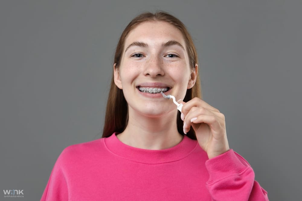 smiling girl metal braces cleaning teeth interdental brush grey background - How to Floss with Braces in Prosper and Colleyville, TX