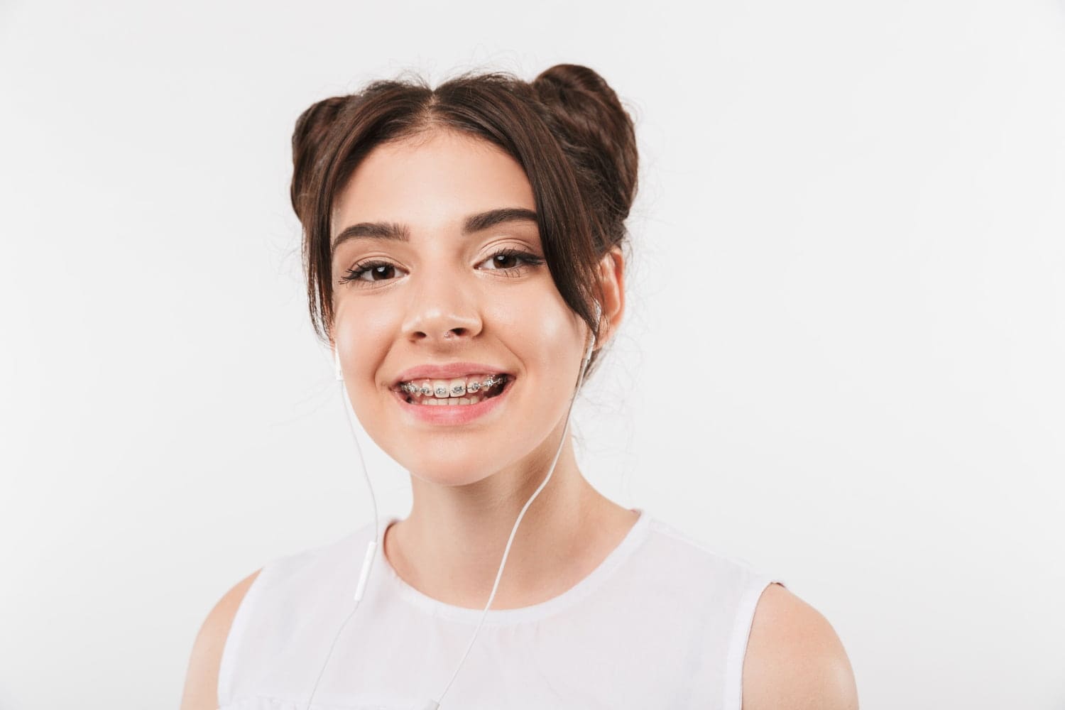 A young woman with brown hair in double buns, white earphones, and braces smiles for Wink Orthodontics in Prosper or Colleyville, TX — showcasing her beautiful smile after learning about Braces Cost.