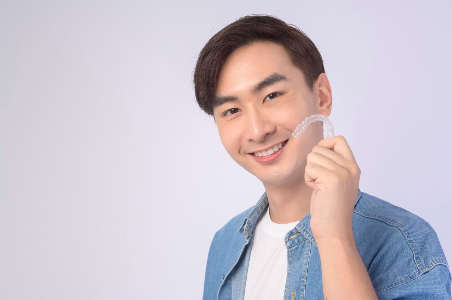 A man in a denim jacket smiles, holding an Invisalign dental aligner for Wink Orthodontics in Prosper or Colleyville, TX.