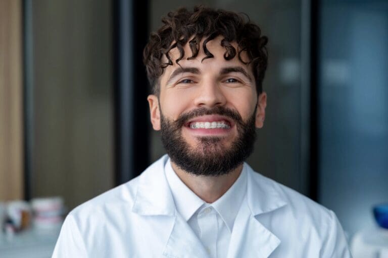 Smiling person with curly hair and a beard wearing a white lab coat, standing indoors - Clear Braces vs Metal Braces in Prosper and Colleyville, TX