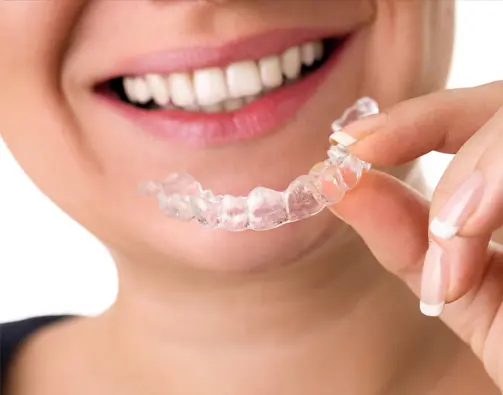 Smiling person with straight white teeth holds a clear aligner from Wink Orthodontics in Prosper or Colleyville, TX.